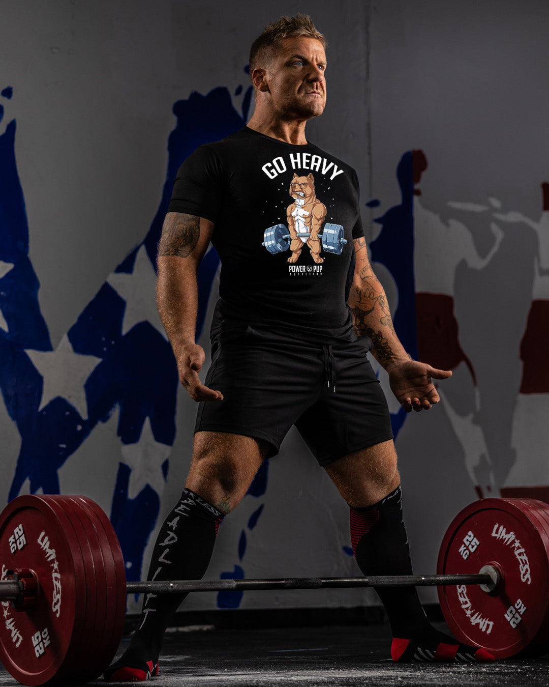 Man lifting weights with a 'Go Heavy' t-shirt in front of a mural.