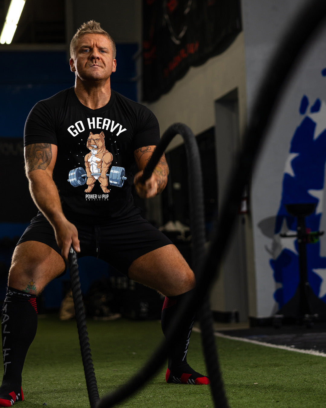 Man in a gym wearing a black t-shirt with 'Go Heavy' graphic, holding battle ropes.