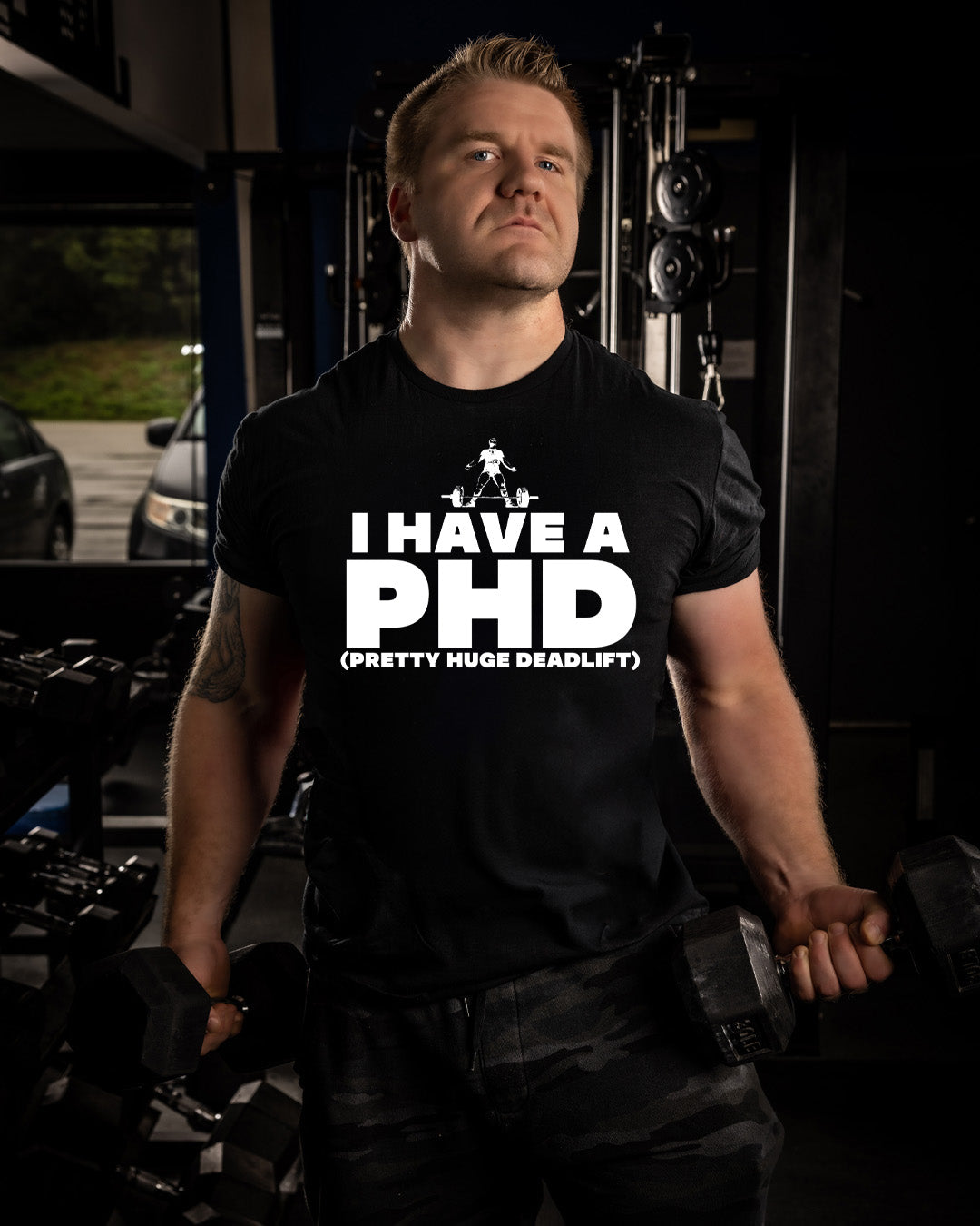 Man wearing a black t-shirt with 'I HAVE A PHD (PRETTY HUGE DEADLIFT)' text in a gym setting.