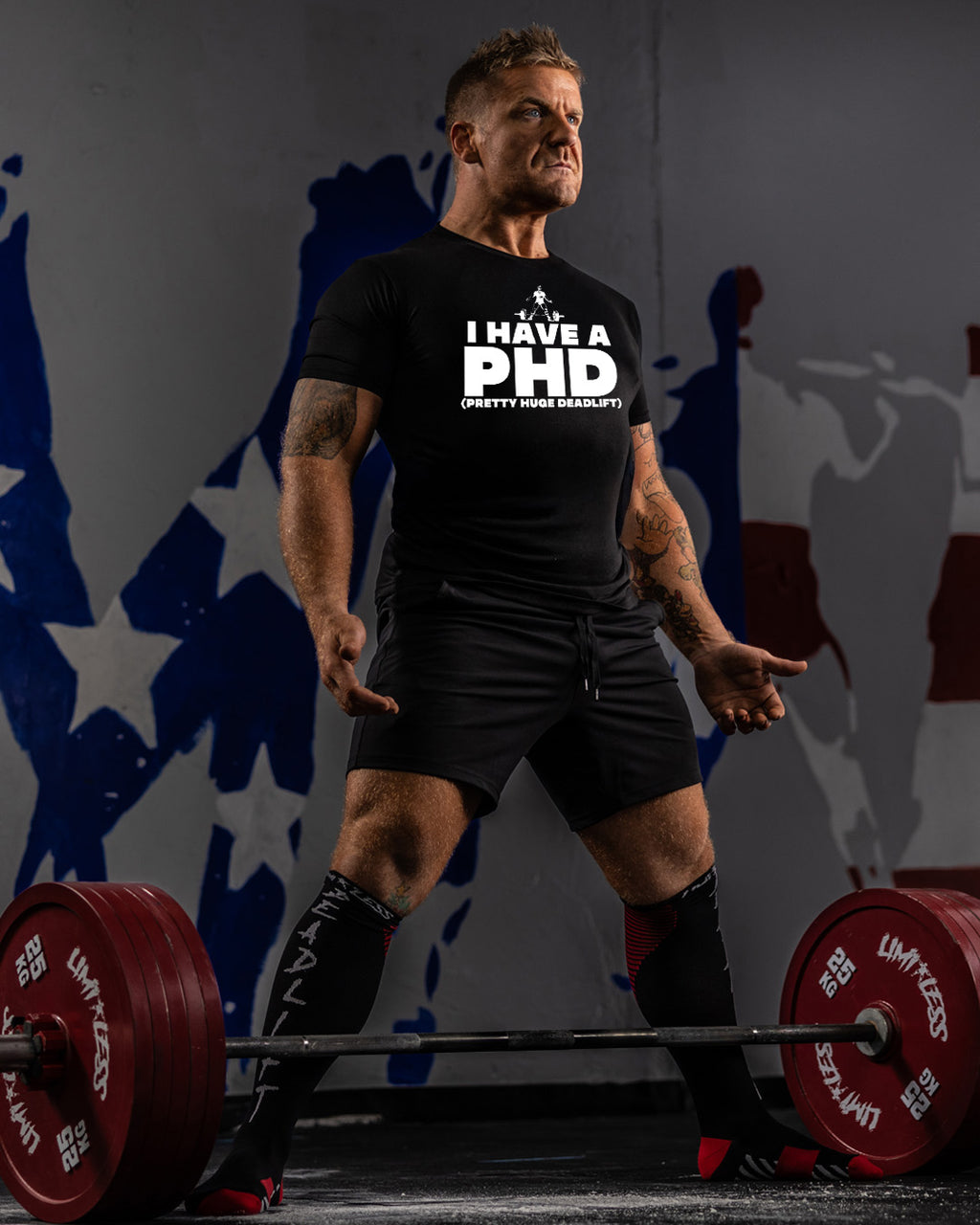 Man lifting weights with a humorous 'I have a PhD' t-shirt in a gym setting