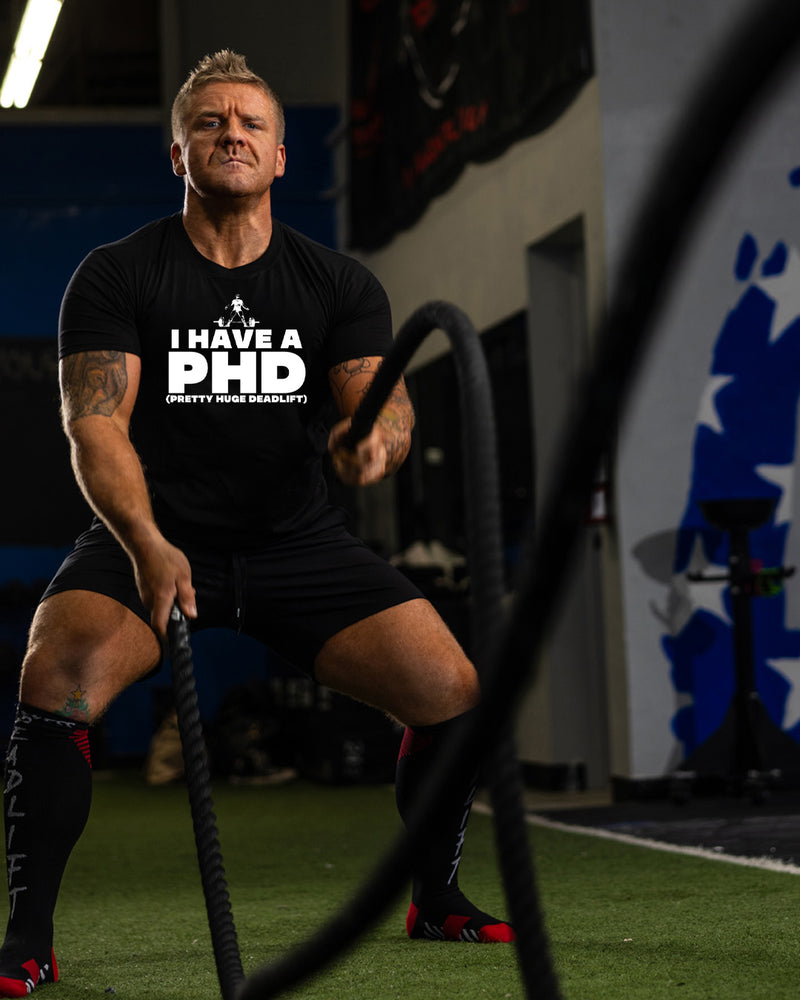 Man in a gym wearing a black t-shirt with 'I Have A PhD (Pretty Huge Deadlift)' text, using battle ropes.