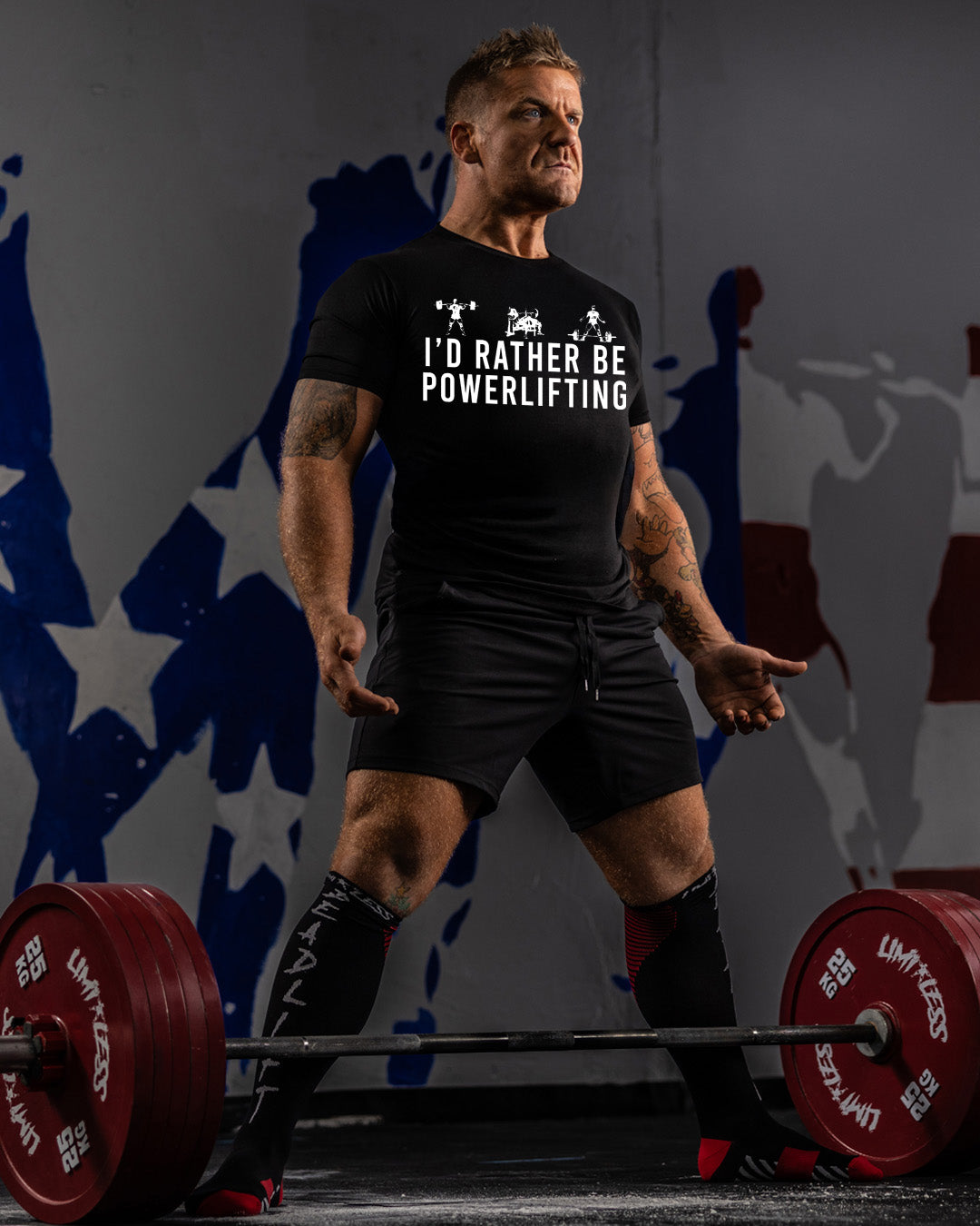Man lifting weights with 'I'd Rather Be Powerlifting' t-shirt against a colorful abstract background