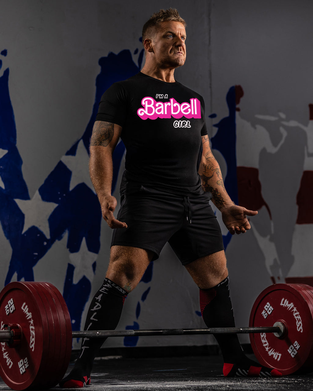 Man lifting weights with a 'Barbell Girl' t-shirt in front of abstract wall art