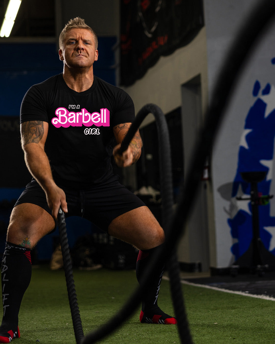 Man in a gym wearing a black t-shirt with 'Barbell Girl' text, holding battle ropes.