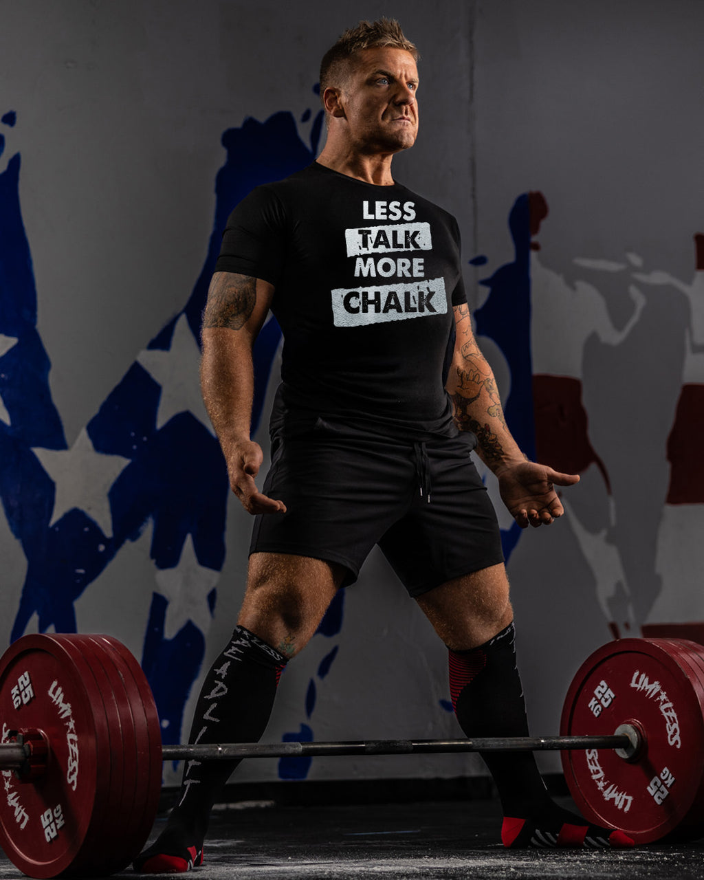 Man lifting weights with a shirt that says 'LESS TALK MORE CHALK' in front of a colorful abstract background.