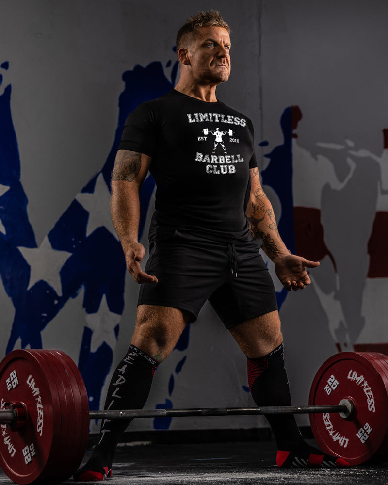 Man lifting weights with a 'Limitless Barbell Club' t-shirt in a gym setting