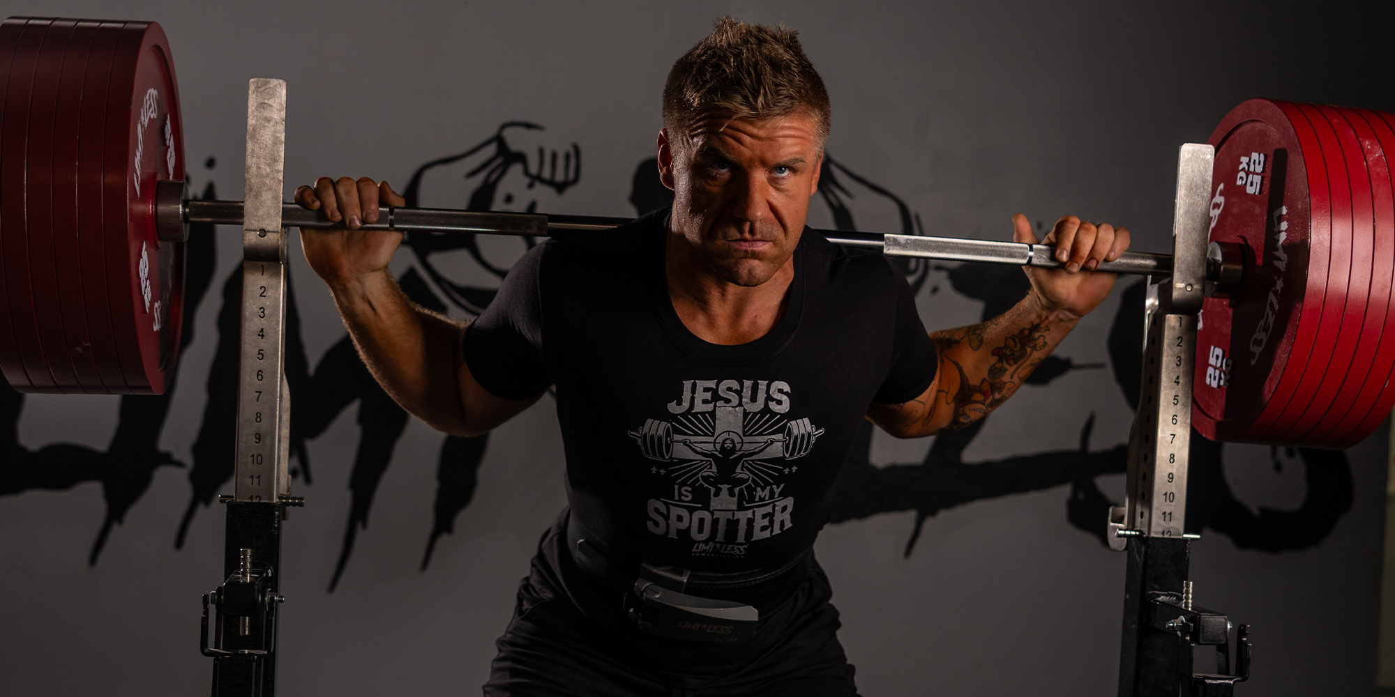 Man lifting weights with a 'Jesus is my spotter' shirt on a dark background