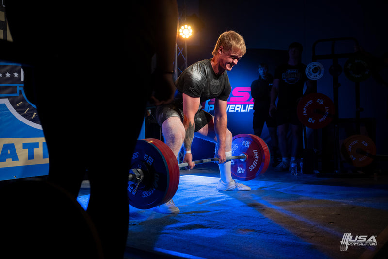 Person lifting a barbell with weights on a stage, likely at a powerlifting competition.