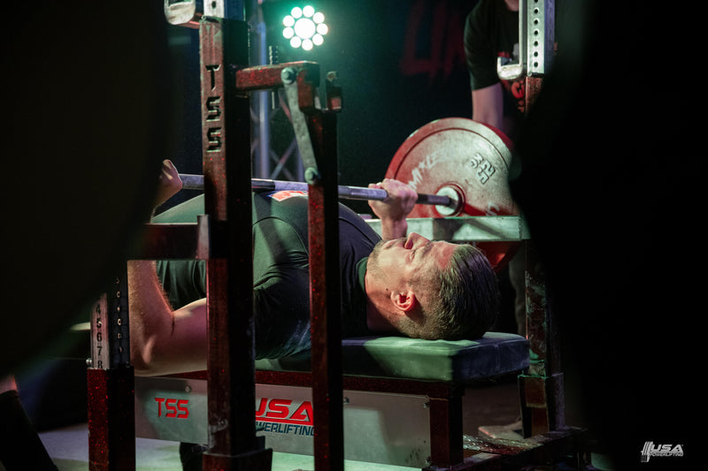 Person performing a bench press with a barbell in a gym setting.  At Limtless Powerlifting USA Powerlifting meet.
