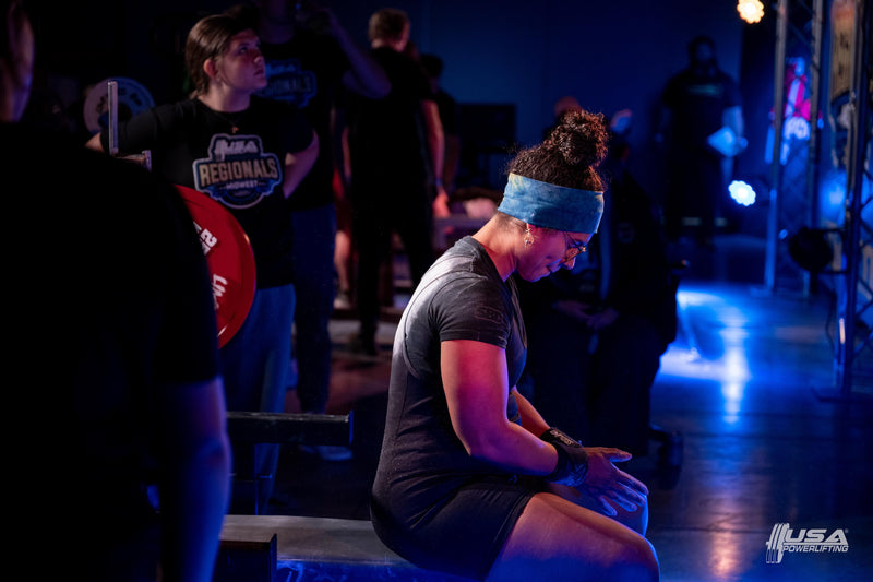 Woman sitting on a bench in a gym setting with blue lighting at USA Powerlifting Midwest Regionals hosted by Limitless Powerlifting