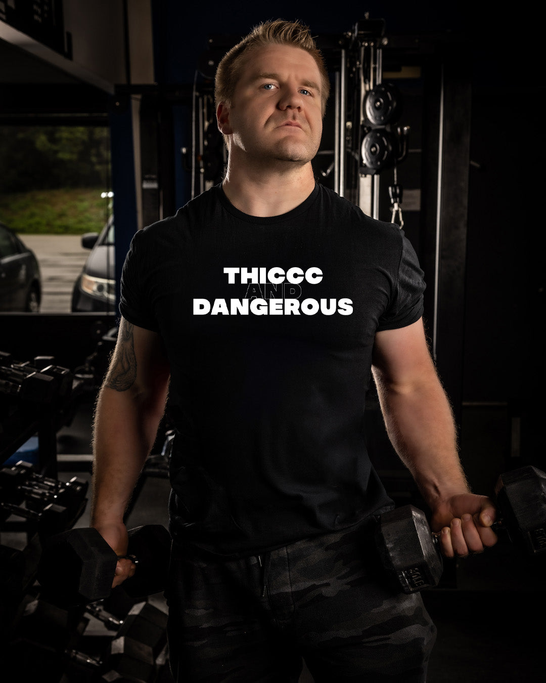 Man wearing a black t-shirt with 'THICC DANGEROUS' text in a dark indoor setting.