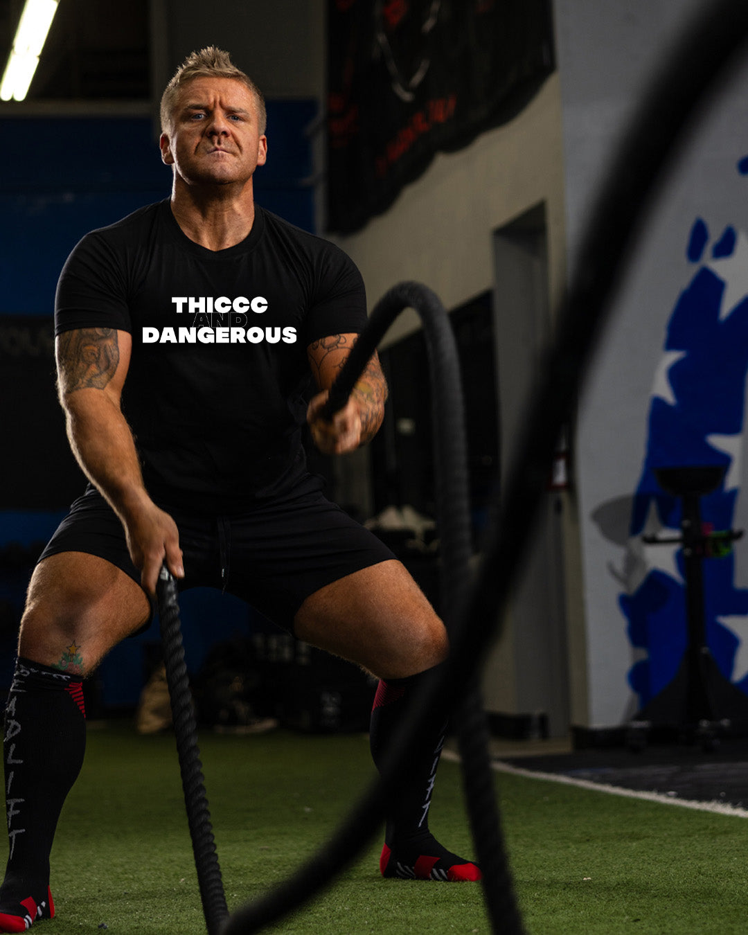 Man in black t-shirt with 'THICC DANGEROUS' text using battle ropes in a gym setting.