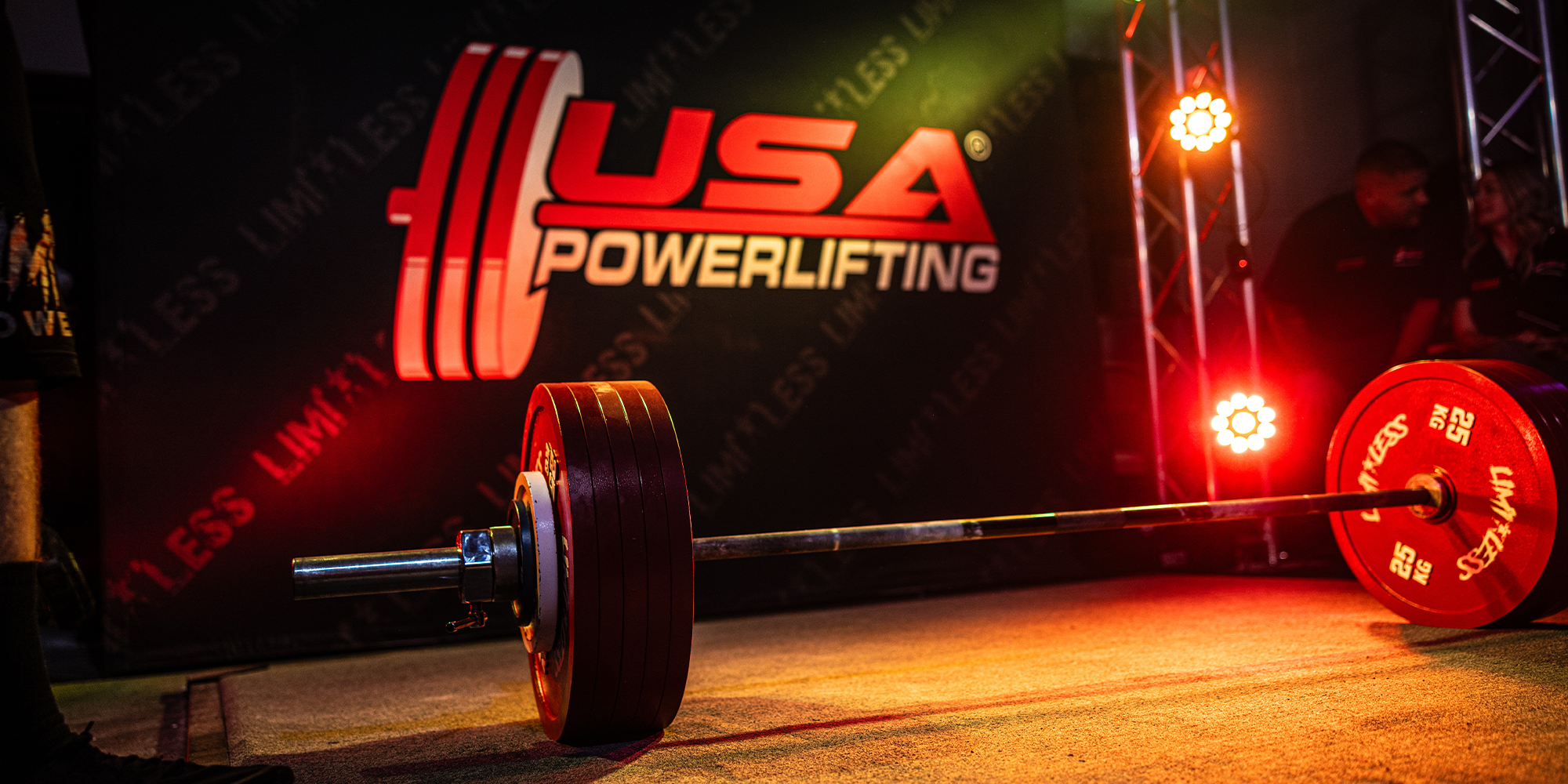 Barbell with Limitless red weight plates on a platform, USA Powerlifting logo in the background