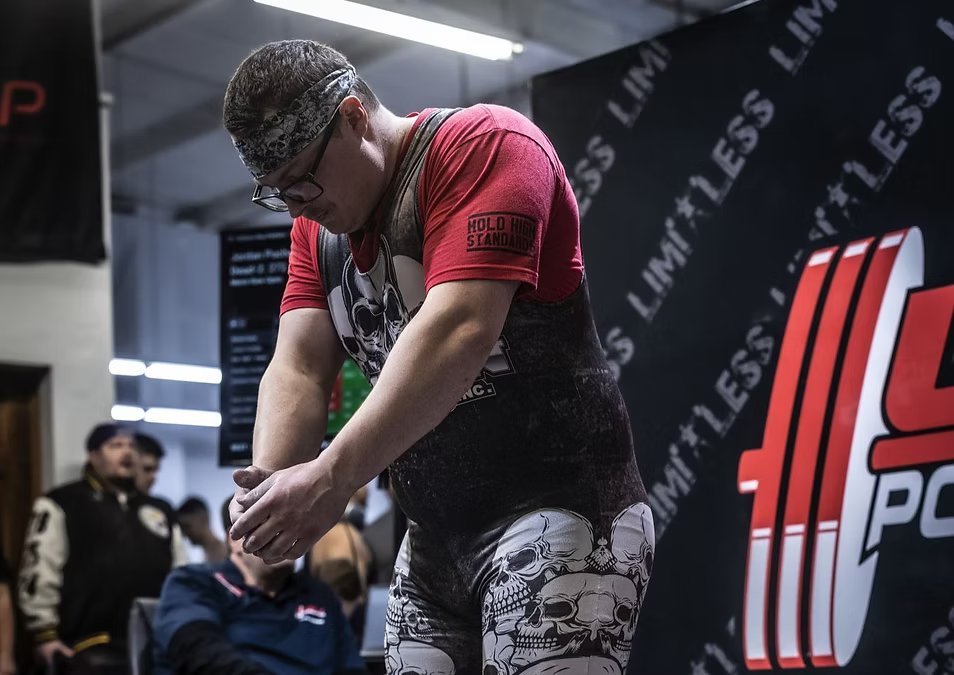 Person in a red shirt and black pants with skull designs, standing in front of a branded backdrop at Limitless powerlifting meet